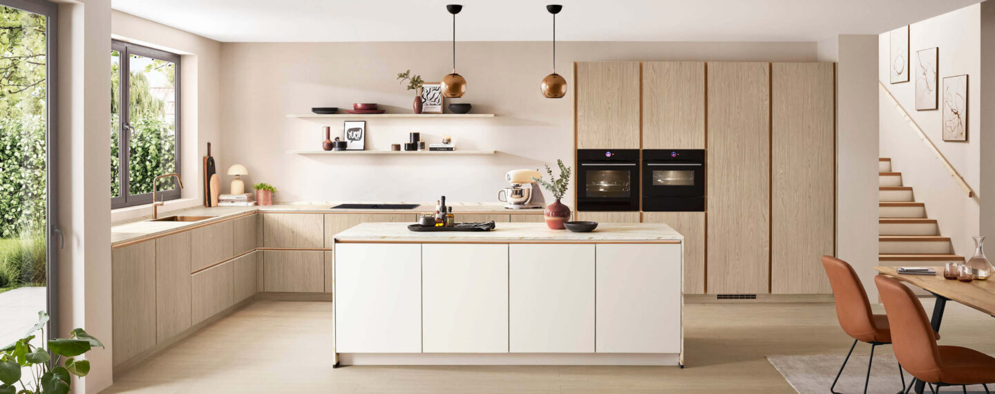 Natürliche Inselküche mit Fronten in Weiß und Holzoptik, goldenen Griffmulden und Arbeitsplatte in Marmoroptik von nobilia erhältlich bei Küchenstudio Ortmann & Winter in Bochum